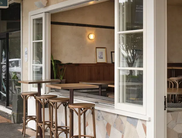 Street-facing facade of a small café or hospitality venue with large white-framed bi-fold service windows opened to an outdoor counter seating area. The exterior combines a pale textured rendered upper wall with a multicoloured stone-clad lower wall, while the interior features timber wall panelling, warm lighting, plants, and café tables and chairs. The image is photographed from the pavement and includes an adjacent glazed entrance.