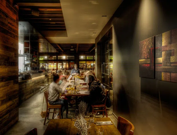 A dimly lit, sophisticated restaurant dining space viewed along a narrow dining room. Guests sit at timber tables with place settings and drinks, surrounded by dark timber wall cladding, exposed timber ceiling beams, glass-fronted shelving, and warm ambient lighting. Abstract geometric artwork decorates the dark wall on the right, creating an intimate, moody atmosphere.