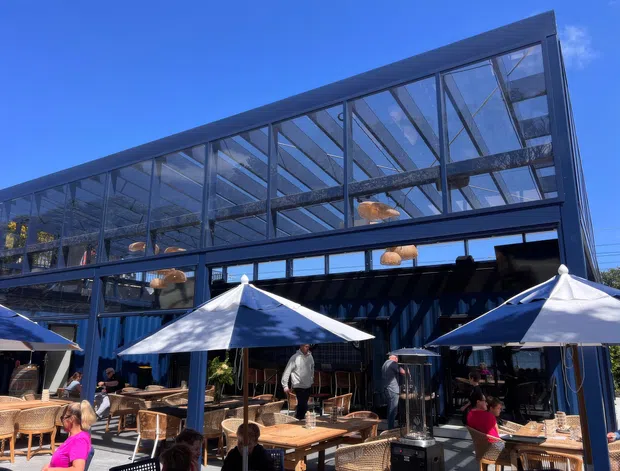 Exterior view of a contemporary multi-level commercial venue with a dark steel-framed glass upper storey and open glazed frontage. The ground-level outdoor dining terrace has white shade umbrellas, timber tables, woven rattan chairs, and patrons seated beneath a clear blue sky.