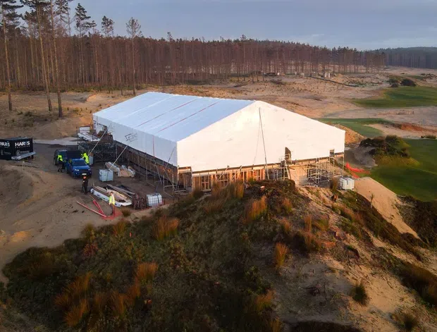 Aerial view of a large temporary or partially constructed rectangular building with a white pitched roof and exposed structural framing, situated on a sandy, sparsely vegetated site surrounded by tall pine trees. Construction equipment, scaffolding and materials are visible around the building. The image appears to document a construction-stage project in a rural or forested landscape.