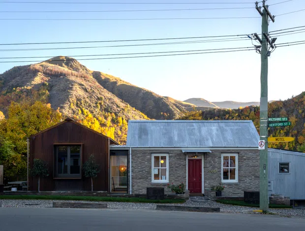 Street-facing view of a compact single-storey building in a mountainous setting. The main structure has a corrugated metal gable roof, grey stone or masonry walls, a red entry door, white-trimmed windows, and a dark timber-clad section on the left. A corrugated metal extension is visible on the right. Utility poles and overhead wires cross the foreground, with autumn-covered mountains rising behind the building.