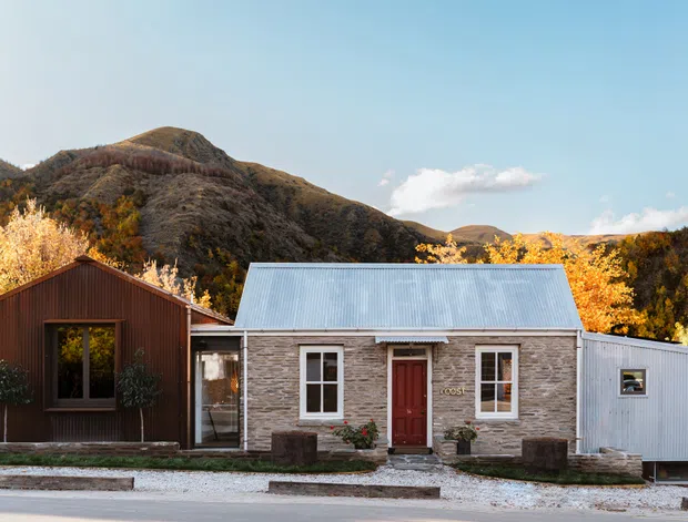 A single-storey residential building in a mountainous autumn setting. The main dwelling has a compact gabled form, a corrugated metal roof, light stone or masonry walls, and a red front door, with a dark timber-clad wing to the left and a corrugated metal-clad extension to the right. The facade faces a paved street and gravel verge, with hills and deciduous trees behind it.