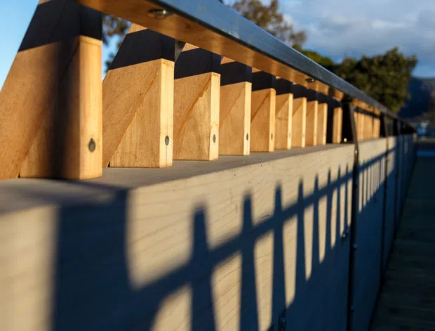 Close-up exterior architectural view of a long timber structure with repeated vertical plywood or timber fins beneath a dark metal roof edge. Strong low-angle sunlight casts rhythmic shadows across the pale wall, while a narrow timber walkway and railing extend alongside the building. The image emphasizes repetitive facade detailing, timber construction, and linear perspective.