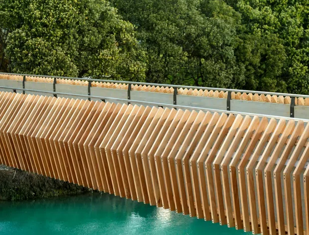 Close-up exterior view of a distinctive pedestrian bridge or bridge-like structure spanning turquoise water. Repeated warm timber ribs form a sculptural side screen or balustrade, with a grey metal upper railing and dense green trees in the background.