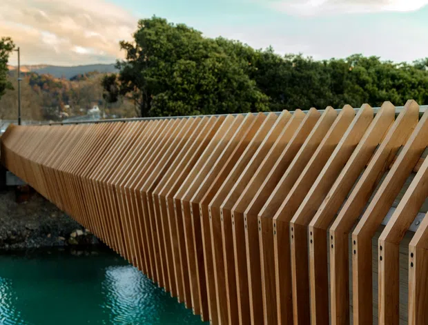 A close view of a distinctive timber pedestrian bridge or elevated walkway with repeated angled slats forming a sculptural balustrade and side structure. The bridge curves over turquoise water, with trees and hills visible in the background.