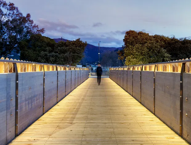A long illuminated pedestrian bridge or elevated walkway with warm timber decking, flanked by symmetrical grey metal or concrete-clad side panels and integrated linear lighting. A lone person walks toward a distant mountainous, tree-covered landscape at dusk.