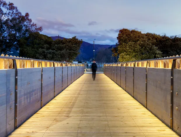 A long illuminated pedestrian bridge or elevated walkway at dusk, with warm timber decking, translucent side panels, and angular illuminated railings. A person walks toward a distant mountainous, tree-lined setting.
