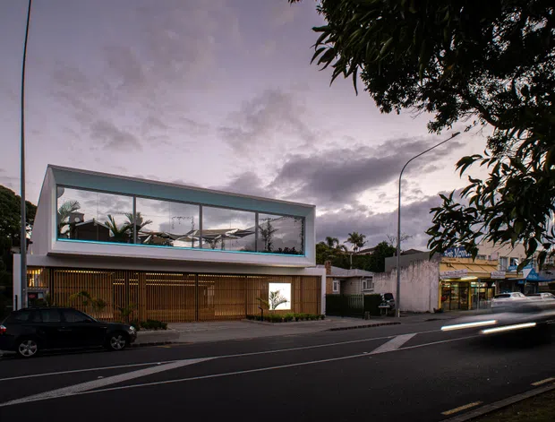 Street-facing evening photograph of a contemporary multi-level building with a strong rectangular glazed upper volume, pale rendered framing, and a screened timber-like lower facade. The building sits on an urban suburban street beside low-rise commercial premises, with palm trees and landscaping visible through the glazing. Vehicle light trails and a dusky sky create a dramatic twilight atmosphere.
