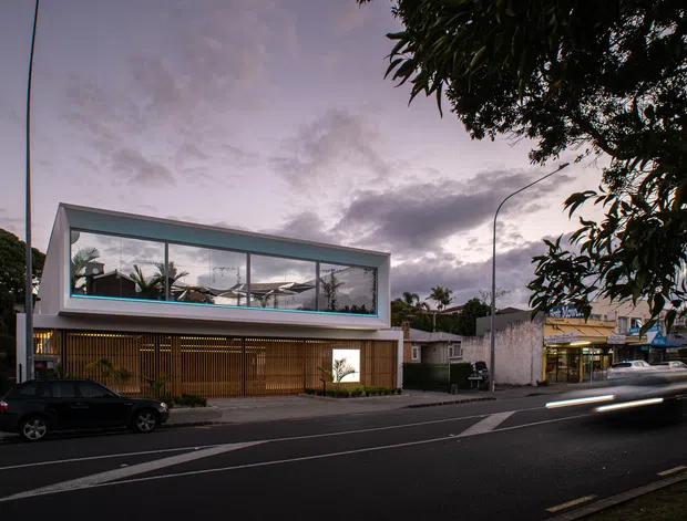 Street-level dusk view of a contemporary two-storey commercial or residential building with a glazed upper volume, projecting flat roofline, and a warm timber screening facade at ground level. The building sits directly along a marked urban roadway with adjacent low-rise buildings and mature street trees.