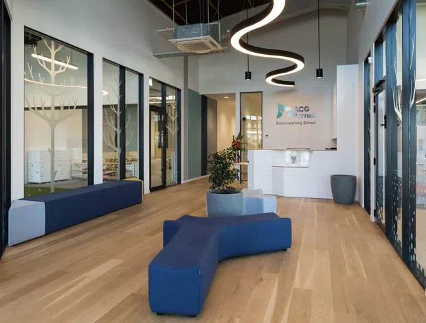 A bright contemporary reception area for an early learning centre, featuring pale timber flooring, white walls, extensive black-framed glazing, a white reception desk, blue curved modular seating, and distinctive sculptural black-and-white pendant lighting. Internal glazed rooms with decorative tree-patterned screening are visible along the left side.