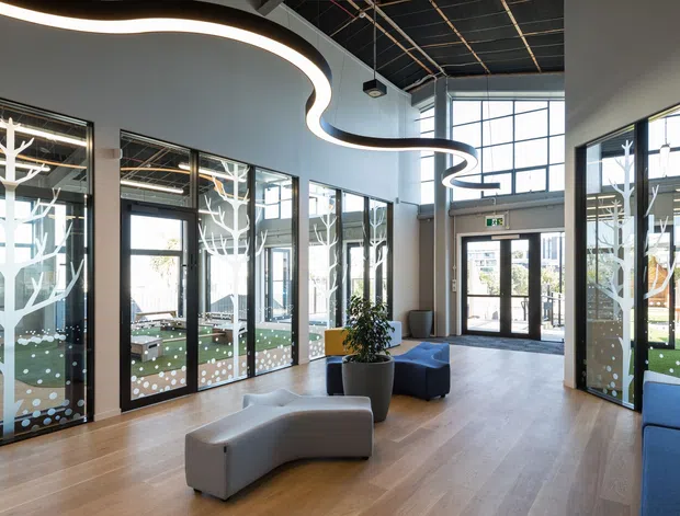 Bright contemporary commercial lobby or entry space with expansive black-framed glazing, light timber flooring, a sculptural curving linear ceiling light, and modern modular seating. Decorative white tree motifs and dot patterns appear on the glass partitions, with a visible outdoor courtyard beyond.