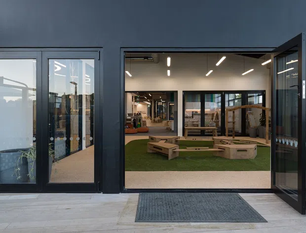 Contemporary commercial or community interior viewed from an exterior threshold, with dark metal-framed glazing and wide folding doors opening onto a bright, open-plan space. The interior features artificial grass, low timber seating and play-like built-in platforms, exposed services, and linear recessed lighting. Pale concrete or stone paving extends across the foreground.
