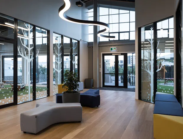 A bright contemporary commercial lobby or entry space with expansive black-framed floor-to-ceiling glazing, pale timber flooring, sculptural curved pendant lighting, and modern modular seating in grey, navy, and yellow. Decorative white tree-like graphics appear on the glazing, with an exterior courtyard and neighboring buildings visible beyond.