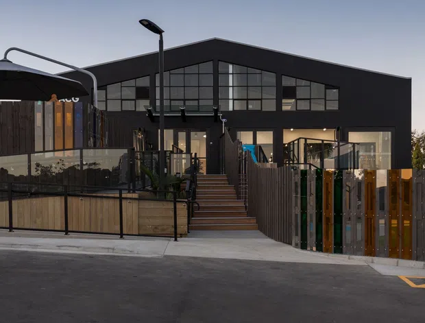 Exterior view of a contemporary dark-clad, multi-level building with a prominent glazed gable facade, rooftop or elevated terrace areas, timber fencing, outdoor umbrellas, and a street-side entrance. A neighbouring residential building is visible in the background.