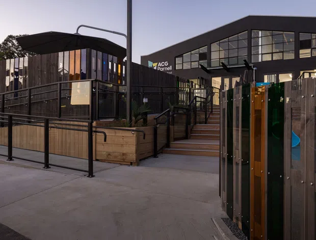 Exterior view of a contemporary commercial building entrance and landscaped access area. The scene features dark timber privacy fencing, black metal balustrades, raised timber planters, stairs, and a modern glazed upper facade with a visible business sign.