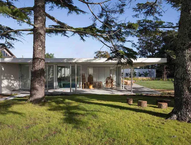 A low, single-storey modern pavilion-style home set within a mature landscaped garden. The building has a broad flat roof, white rendered walls, extensive floor-to-ceiling glazing, and an open living area with timber furniture visible through the glass. Large established trees frame the foreground, while a paved terrace and lawn extend around the house.
