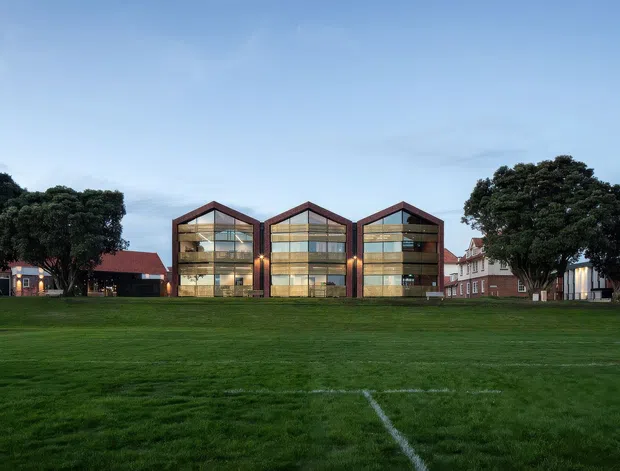 Wide exterior view of a contemporary multi-storey building composed of three adjoining gabled volumes, with extensive floor-to-ceiling glazing, dark structural framing, and warm timber-toned facade elements. The building fronts a broad manicured grass field and is surrounded by mature trees and nearby traditional pitched-roof buildings under an overcast sky.