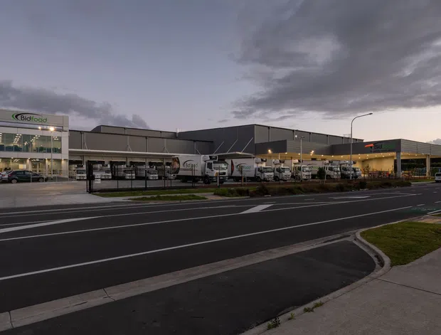 Exterior twilight view of a large contemporary commercial and industrial facility, with a dark metal-clad warehouse or distribution building, glazed office entrance, loading and delivery vehicles, landscaped frontage, and a marked roadway in the foreground.