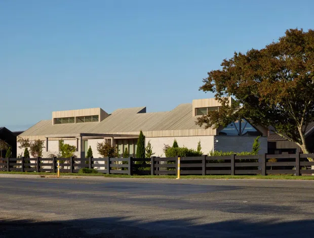 Street-facing view of a low-rise contemporary building with a broad pale metal pitched roof, raised clerestory-like roof sections, dark timber-style fencing, landscaped shrubs, and mature trees. The building appears to have a mixed residential or small commercial character, with the roof and facade dominating the composition.