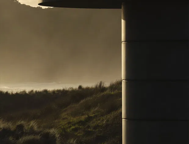Moody architectural detail of a dark cylindrical building element or tower with a projecting curved canopy, silhouetted against a windswept coastal landscape of dunes and long grass. Warm low-angle sunlight, hazy sky, and strong shadow create a dramatic, minimalist composition. No interior is visible.