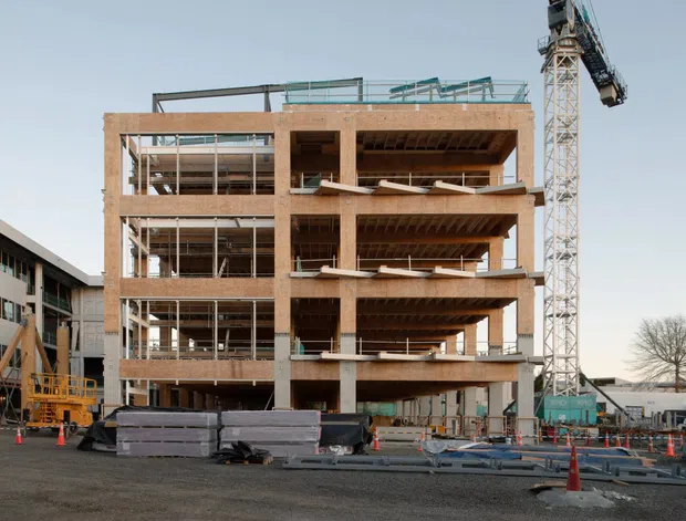 Exterior photograph of a multi-storey building under construction, showing exposed concrete floor slabs, structural columns, open floor plates, brick-toned facade elements, temporary safety barriers, construction materials, and a tower crane. The surrounding site and adjacent construction structures reinforce the active building-site context.