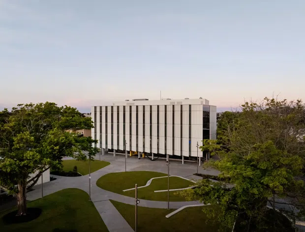 Exterior architectural photograph of a modern multi-storey institutional or commercial building set within landscaped grounds. The building has a rectilinear form, a flat roof, a pale facade with repeated narrow vertical window bays or fins, and a darker glazed end wall. Curving pedestrian paths, lawns, mature trees, and slender site light poles surround the building.