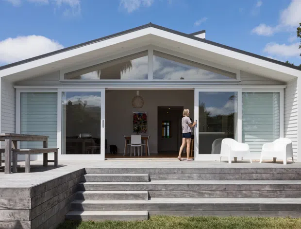 Front exterior view of a single-storey contemporary coastal-style home with a broad gabled roof, white horizontal cladding, extensive glazed sliding doors, and a raised timber deck accessed by wide steps. The open interior is visible through the glazing, with a small dining or living area and people near the entry. Outdoor seating and a picnic-style table occupy the deck.