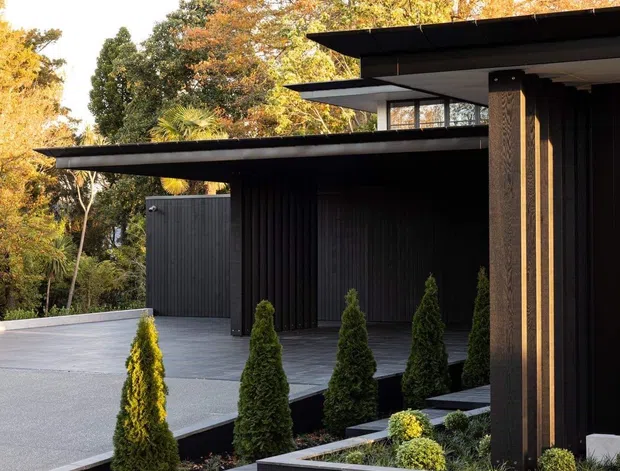 A contemporary dark-clad residential exterior with strong horizontal flat roof planes, deep eaves, and covered entry or carport-like volumes. The building is set within a carefully designed landscape featuring dark paved paths, large grey paving, raised black planter boxes, and regularly spaced conical shrubs. Warm autumn trees form the background, contrasting with the charcoal architecture.