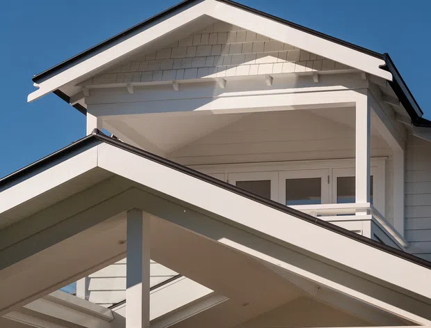 Close-up exterior view of a multi-level white gable-roofed house, showing overlapping pitched roof forms, covered balcony and terrace structures, exposed gable detailing, white weatherboard cladding, dark roof edging, and glazed doors. The image is photographed against a clear blue sky with strong sunlight and deep architectural shadows.