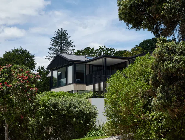 Exterior view of a contemporary elevated house partially screened by mature landscaping. The home has dark metal pitched roof planes, extensive glazing, dark vertical framing and railings, and a raised light-coloured lower wall above a rough stone retaining edge. A sandy unsealed foreground and abundant coastal-style vegetation surround the property under a bright blue sky.
