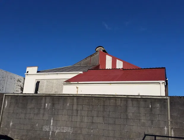 Exterior view of a compact older building behind a tall concrete block boundary wall. The structure has a prominent red metal hipped roof with a raised central section, white rendered walls, and a small chimney or vent. Snow-dusted mountainous terrain is visible in the background beneath a clear blue sky.