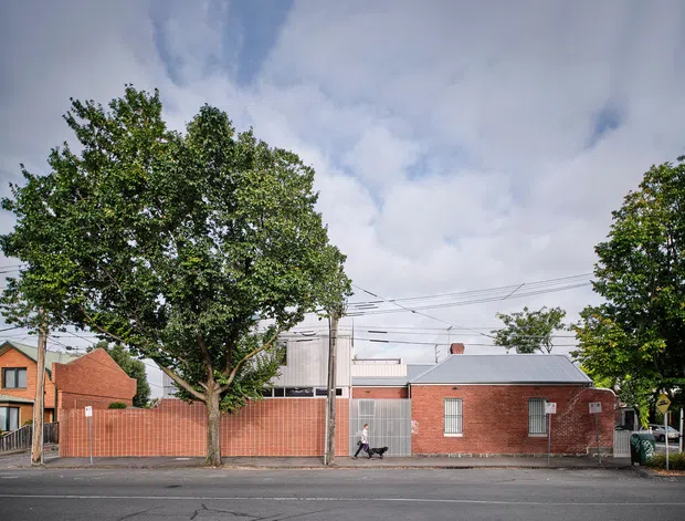Street-level photograph of a low-rise urban brick building behind a red brick boundary wall, with a corrugated metal roof, narrow barred windows, utility wires, mature street trees, and neighbouring brick homes. The image primarily documents the building facade and its streetscape context.