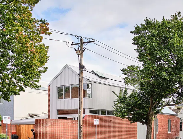 Street-level exterior view of a contemporary multi-storey urban residence, with a corrugated light-grey upper facade, red brick boundary wall and adjoining brick structure. A sloped roof with a prominent skylight, large windows, timber gate, street trees and overhead utility wires are visible.