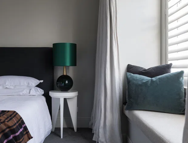 A compact, contemporary bedroom corner with a partially visible bed, dark upholstered headboard, white bedding, and a small white geometric bedside table. A dark green table lamp sits beside the bed, while a deep window seat with blue-grey cushions occupies the right side beneath white plantation shutters. Soft grey curtains and neutral finishes create a calm, refined atmosphere.