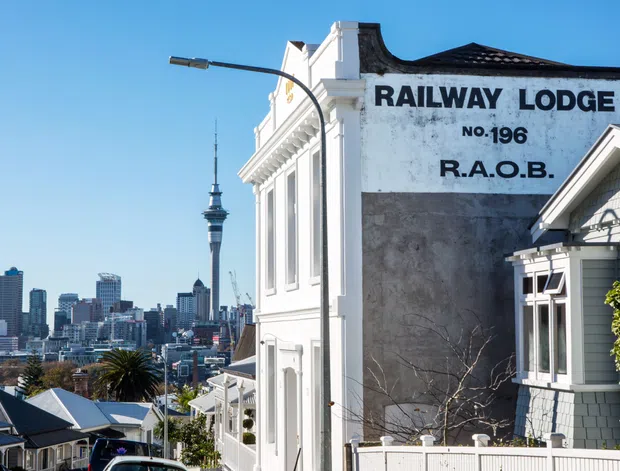 Exterior streetscape view of a historic white-painted lodge building on a sloping urban site, with ornate classical detailing, traditional window surrounds, a tiled roof, and prominent painted signage reading “RAILWAY LODGE NO.196 R.A.O.B.” A dense city skyline and tall tower are visible in the background.
