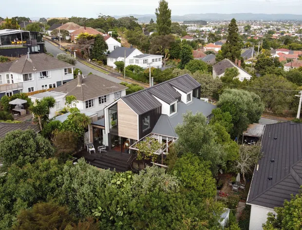 Aerial view of a leafy suburban neighbourhood on a hilly site, centred on a contemporary multi-level house with dark grey pitched roof forms, timber-toned cladding, and extensive glazing. Surrounding homes, mature vegetation, streets, and a broad urban landscape are visible, with distant hills on the horizon.