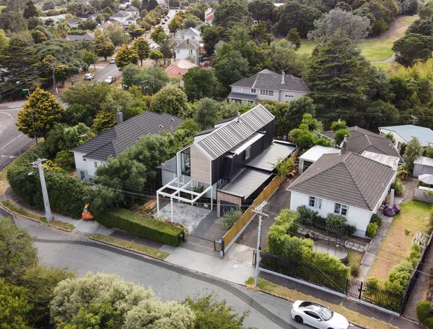 Aerial view of a suburban residential neighbourhood with a contemporary multi-level home prominently positioned among established houses, gardens, tree-lined streets and gently sloping terrain. The featured dwelling has a dark, angular pitched roof, light cladding, extensive glazing, a raised terrace or balcony, and a compact landscaped frontage.