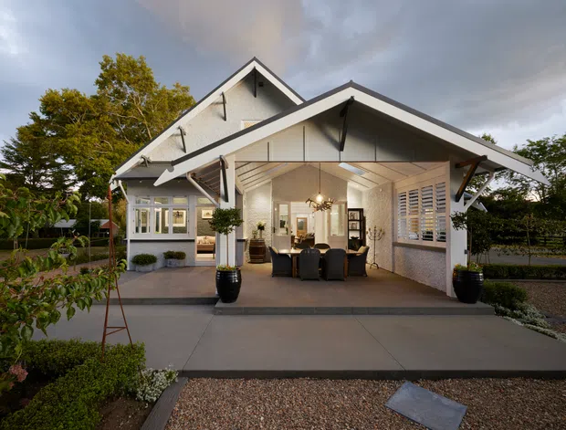 Exterior view of a detached, gabled home with a broad covered outdoor living area opening into a bright dining space. The building has light painted weatherboard-style cladding, dark trim, exposed roof structure and decorative brackets, large glazed openings, and landscaped gravel and planting around a concrete forecourt. The image has a refined traditional farmhouse character with a warm, inviting atmosphere.