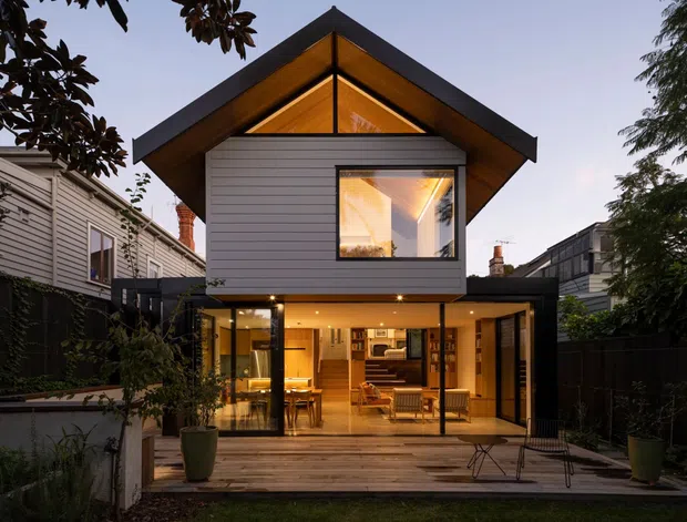Exterior view of a compact contemporary two-storey house in an established residential setting. The home features a prominent steep gable roof with dark metal edging, warm timber lining beneath the eaves and in the upper gable, horizontal light-grey weatherboard cladding, and extensive ground-floor glazing opening onto a timber deck. The illuminated interior reveals a kitchen and dining/living area, while mature planting and neighbouring homes frame the property.