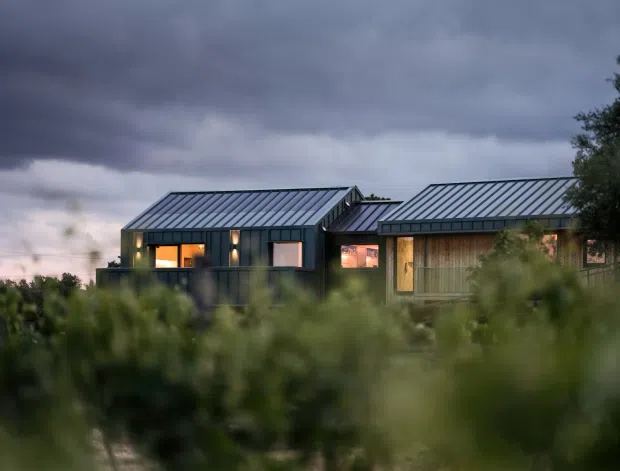 A low, single-storey modern residence photographed at dusk through blurred vegetation. The house is composed of connected rectangular volumes with dark vertical cladding, standing-seam metal gable roofs, large windows glowing with warm interior light, and a timber-clad section on the right. Heavy storm clouds create a dramatic rural landscape setting.