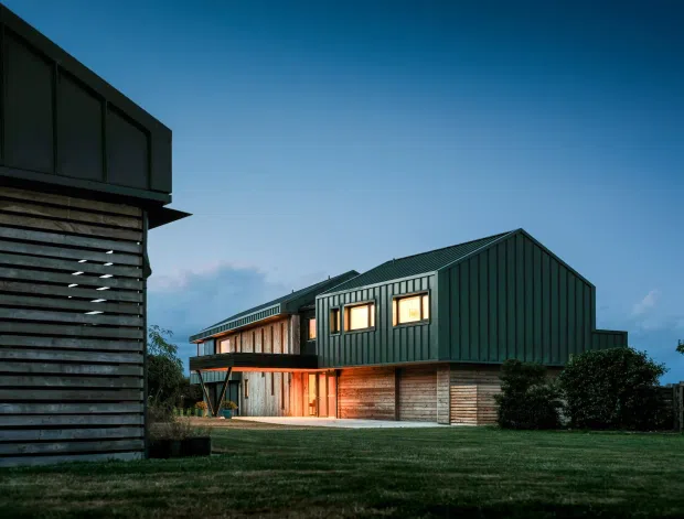 Exterior architectural photograph of a contemporary multi-volume house at dusk. The building features dark green vertical metal cladding and pitched gabled roofs, with lower walls clad in horizontal timber weatherboards. Warm light glows from the windows and recessed entry, while a slatted timber-clad structure frames the left foreground. The house sits in a landscaped grassy setting.