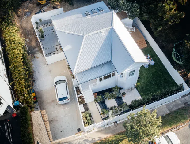 Aerial view of a compact light-coloured residential house with a prominent pitched metal roof, several roof skylights, white exterior walls, a covered front entry, landscaped garden, paved driveway and boundary fencing. The property appears to be in a suburban setting and includes a raised timber deck at the rear.