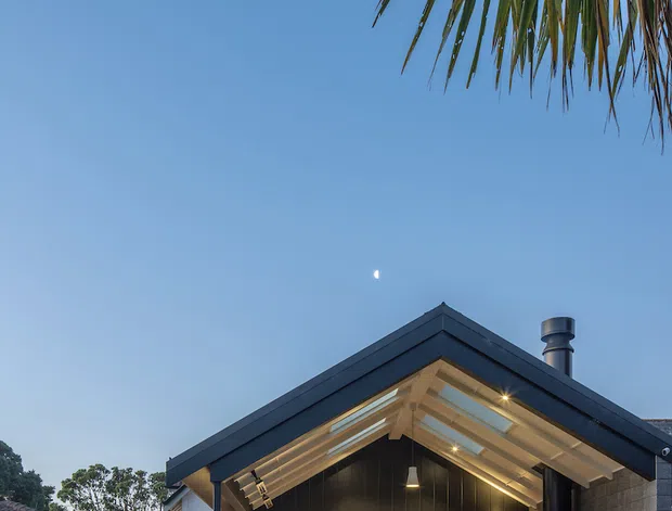 Exterior view of a contemporary gable-roof extension with a covered timber deck and outdoor entertaining area. The dark charcoal-clad gable facade contrasts with pale exposed roof lining, masonry side walls, and warm interior lighting visible through large glazed doors. The home sits in a suburban garden setting, with neighbouring rooflines, lawn, mature trees, and palm foliage visible.