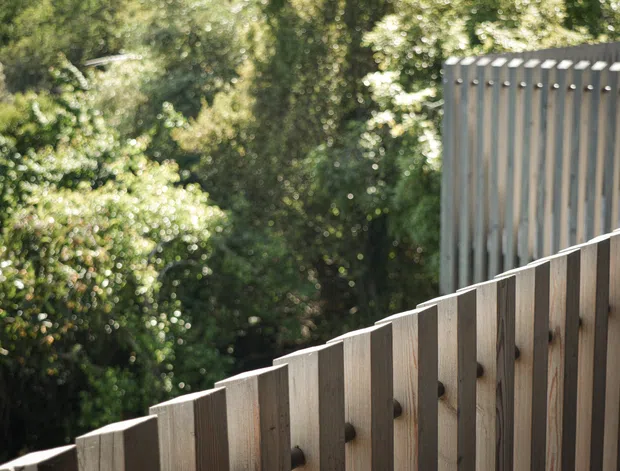 Close-up exterior architectural detail showing a slatted timber fence or balustrade rising diagonally across the foreground, with a taller grey vertical-slatted screen or facade element behind it. Dense leafy vegetation forms the background, and sunlight highlights the natural wood grain and repeating vertical rhythm.