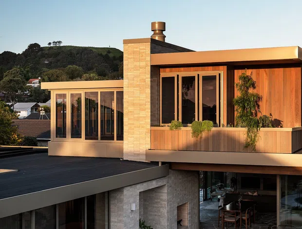 Exterior architectural photograph of a contemporary multi-level residence on a sloping suburban site. The house features strong horizontal flat rooflines, extensive glazing, warm timber-clad upper-level walls, pale masonry and stone finishes, a prominent chimney, and a sheltered outdoor dining or living area opening onto irregular stone paving and landscaped planting. Hillside vegetation and neighbouring homes are visible in the background.
