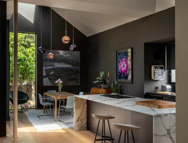 A sophisticated open-plan interior combining a kitchen and dining area. The space features warm timber flooring, dark charcoal walls, a large white-and-grey veined marble kitchen island with waterfall ends, dark cabinetry, timber dining furniture, sculptural pendant lights, and floor-to-ceiling glazing opening toward greenery. A compact coffee or service nook is visible at the rear right. The high, angular ceiling and strong natural light create a dramatic yet refined atmosphere.