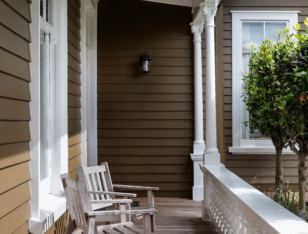 A covered traditional front veranda with weatherboard walls, white decorative columns and balustrade, timber decking, tall white-trimmed windows, and two wooden outdoor chairs. The space has a refined heritage character with warm brown cladding and abundant natural light.
