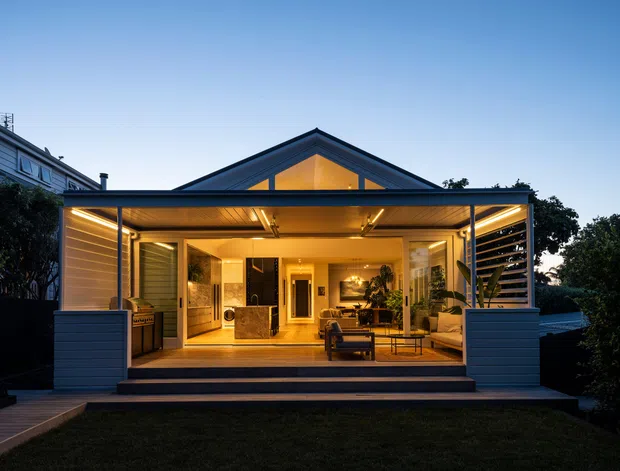 Twilight exterior view of a contemporary single-storey house extension opening onto a covered outdoor living area. The facade features dark blue-grey horizontal cladding, extensive glazed sliding doors, a pitched gable roof with a warm illuminated triangular clerestory, and timber decking with broad steps. The covered space contains a kitchen and lounge/dining area, creating a strong indoor-outdoor connection.
