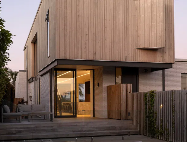 Exterior view of a contemporary multi-storey residence at dusk, featuring a tall gabled timber-clad upper volume above a light brick lower level. Large glazed doors open onto a raised timber deck with outdoor seating, while a swimming pool occupies the foreground. Vertical timber fencing and subdued integrated lighting frame the private courtyard setting.