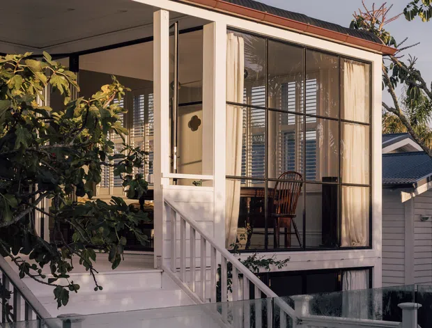 Exterior view of a raised, light-painted timber house with a gabled roof and corrugated dark roofing. The facade features a covered porch, enclosed sunroom or living area with large black-framed glazing and curtains, white balustrades, stairs, and a glass-and-white fence. Mature planting partially screens the front elevation.